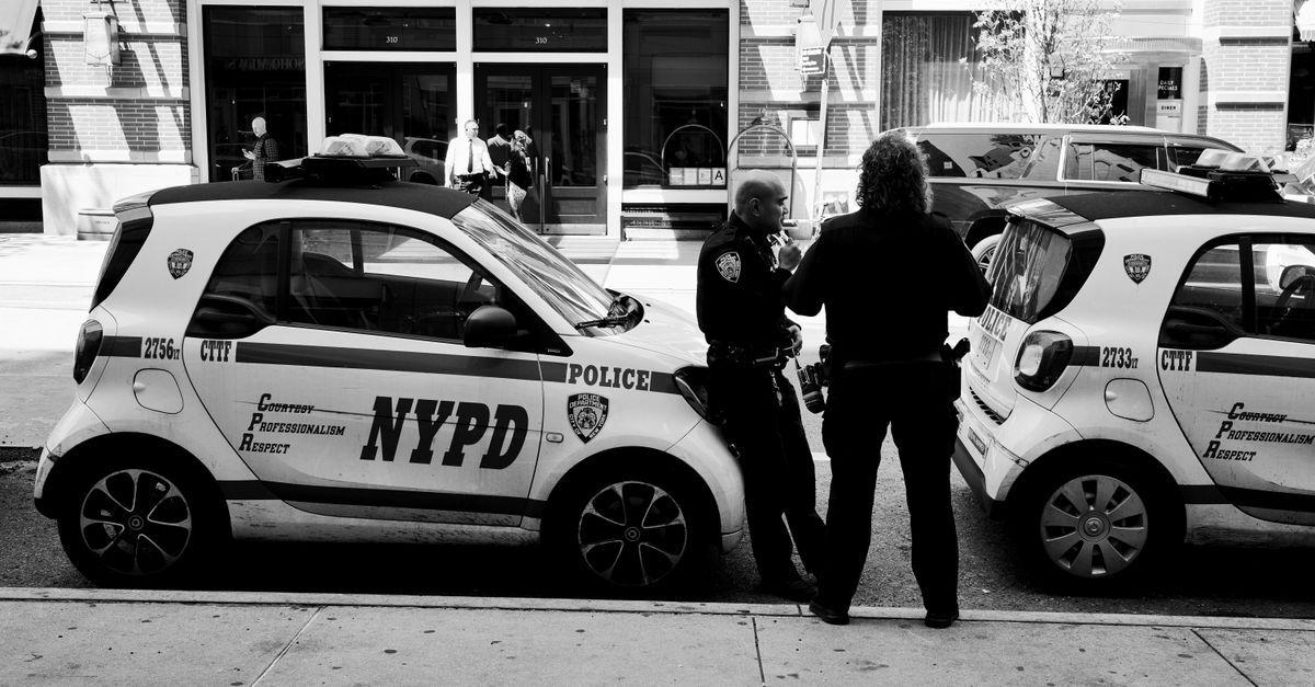 NYPD officers in conversation beside marked police cars on a city street, captured in black and white.